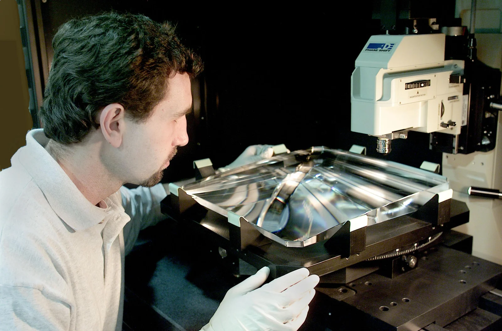A worker wearing white gloves inspects a large rectangular focusing lens beneath measurement equipment.
